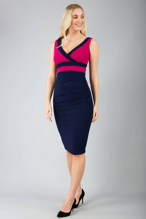 A tall blonde woman stands face on in a form fitting bodycon dress with a raspberry coloured bodice and navy skirt. navy contrast bands are at the bust to emphasise the crossover , and there is ruching across the front of the tummy area.