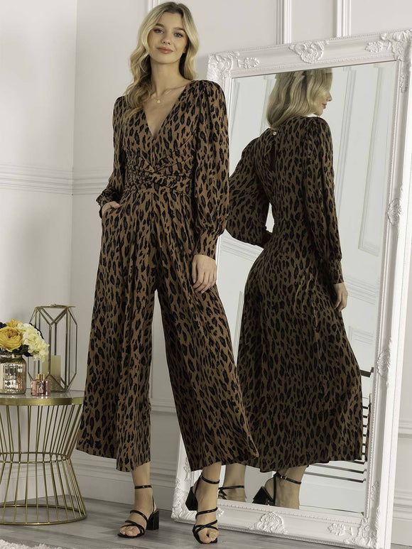 A tall slim blonde woman wearing a wide leg jumpsuit in a brown leopard print. it has cuffed blouson sleeves and a crossover neckline, with a ruched waist, plus one hand is in a side pocket.