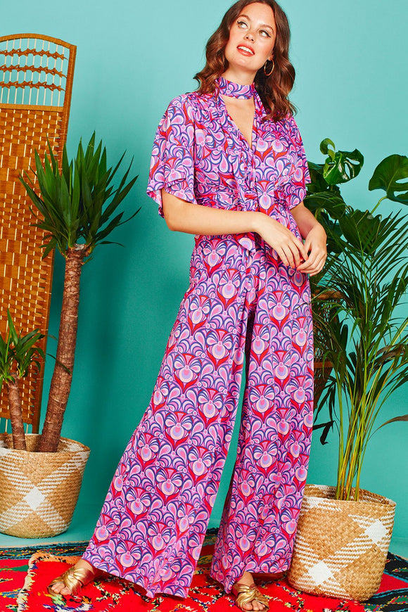 A tall brunette woman poses in a fabulously flowing, boldly patterned pink and purple jumpsuit, with a matching tie at her neck. The legs are wide and there is a gentle v neck, and the material has a gentle sheen.