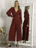 A tall woman with hand on hip facing the camera, wearing a red animal print jumpsuit that has long blouson sleeves and wide palazzo legs. there are flattering gathers in a v shape at the waist, and the neckline is an elegant plunging v.