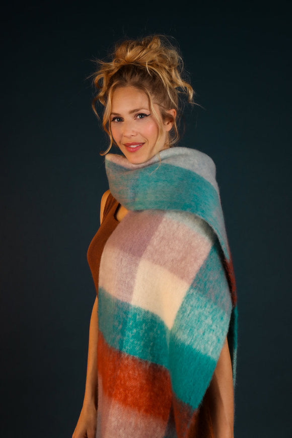 Maddie Cosy Scarf - a tall blonde woman wears a huge slightly fluffy looking scarf that has large squares of plush autumn jewel tones over it. The scarf wraps over her shoulder but easily covers her torso on each side too. 