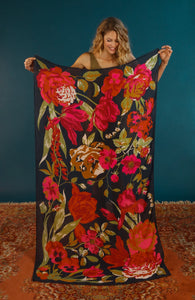  Floral Tapestry Scarf by Powder. Huge punchy florals in rich tones on a navy background on a huge rectangular scarf held up by a blonde woman. 