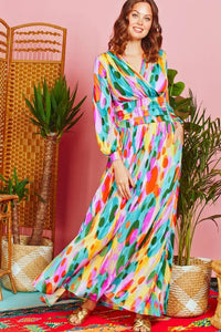 A tall brunette woman wears a crossover bust maxi dress with gathered waist and a flowing skirt, topped with pockets and long bishop sleeves. the dress is covered in gorgeous large spots of varying sizes in shades of green, yellow, orange and pink. It is a silk jersey fabric maxi by Onjenu.